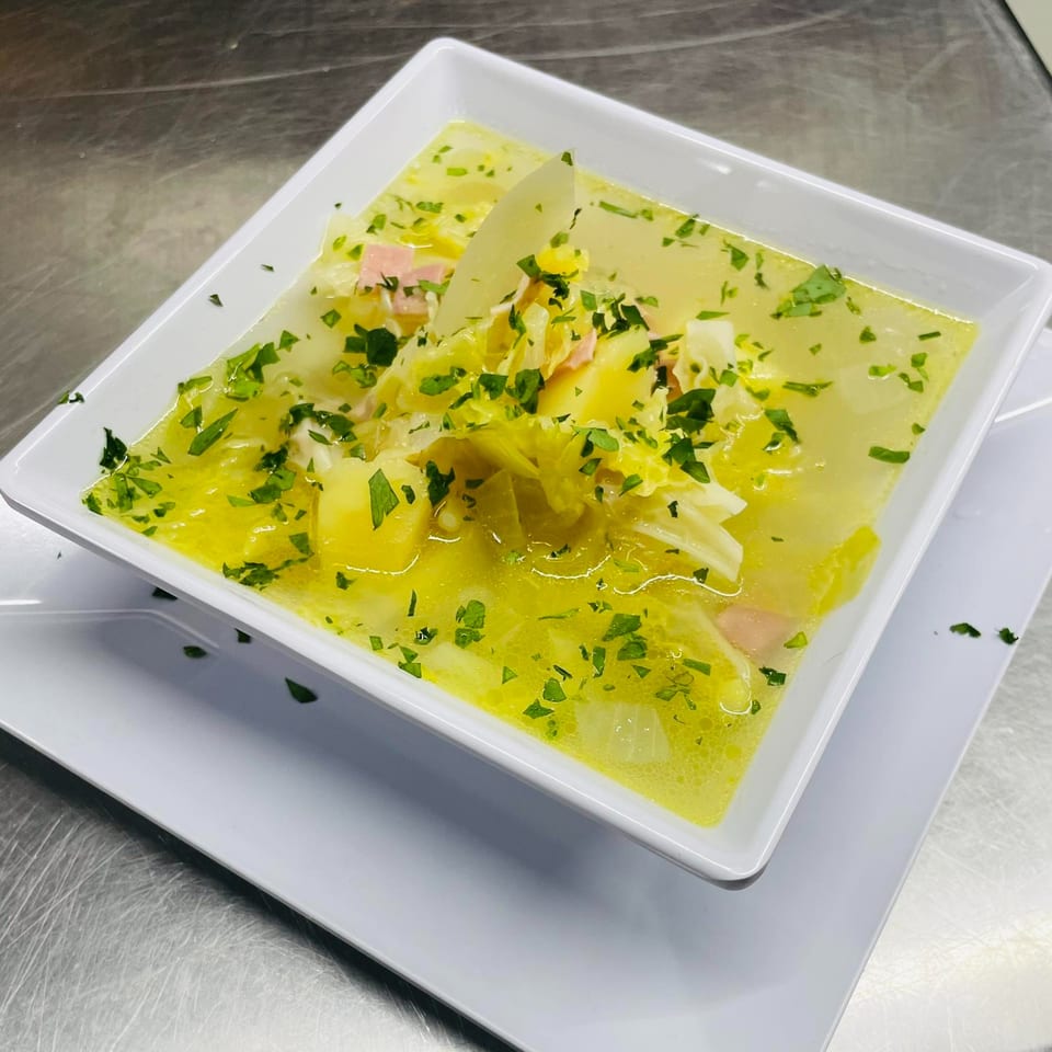 A white square bowl of hearty soup with cabbage, potatoes, ham, and fresh parsley.
